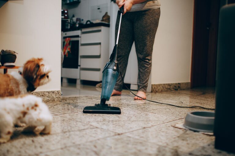 A woman cleaning the floor with a vacuum cleaner while a dog watches attentively. Indoor home setting.
